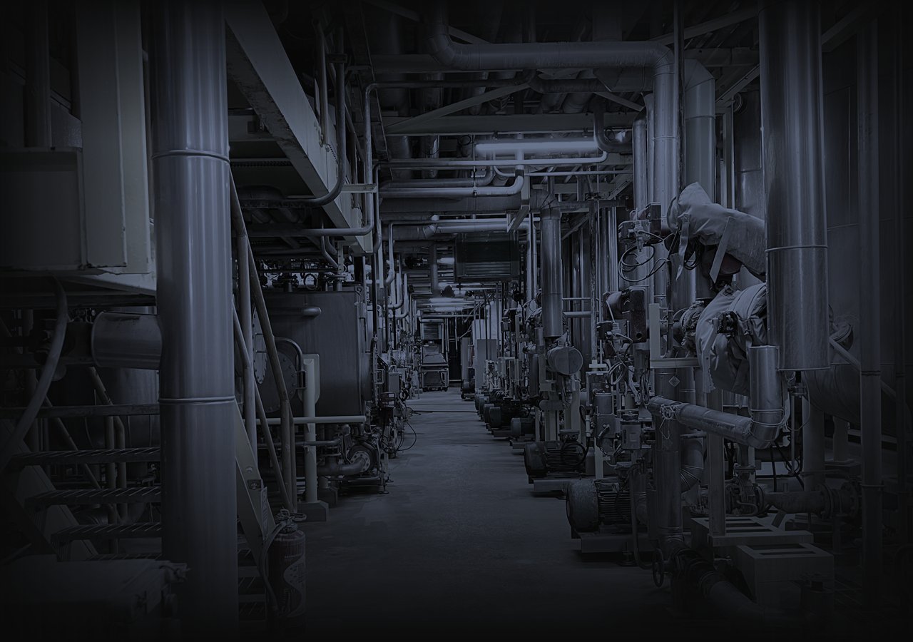 Service alley in a plant, pipework and pumps lined up on both sides under fluorescent light, graded in cold grey for the visual essay The grey zone.