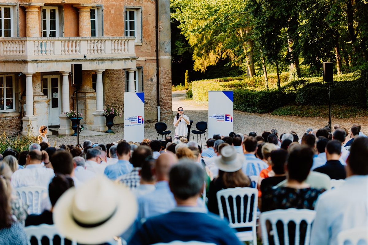 Asma Mhalla at the microphone addressing an audience of executives, outdoors in front of a manor, at the MEDEF Somme garden party.